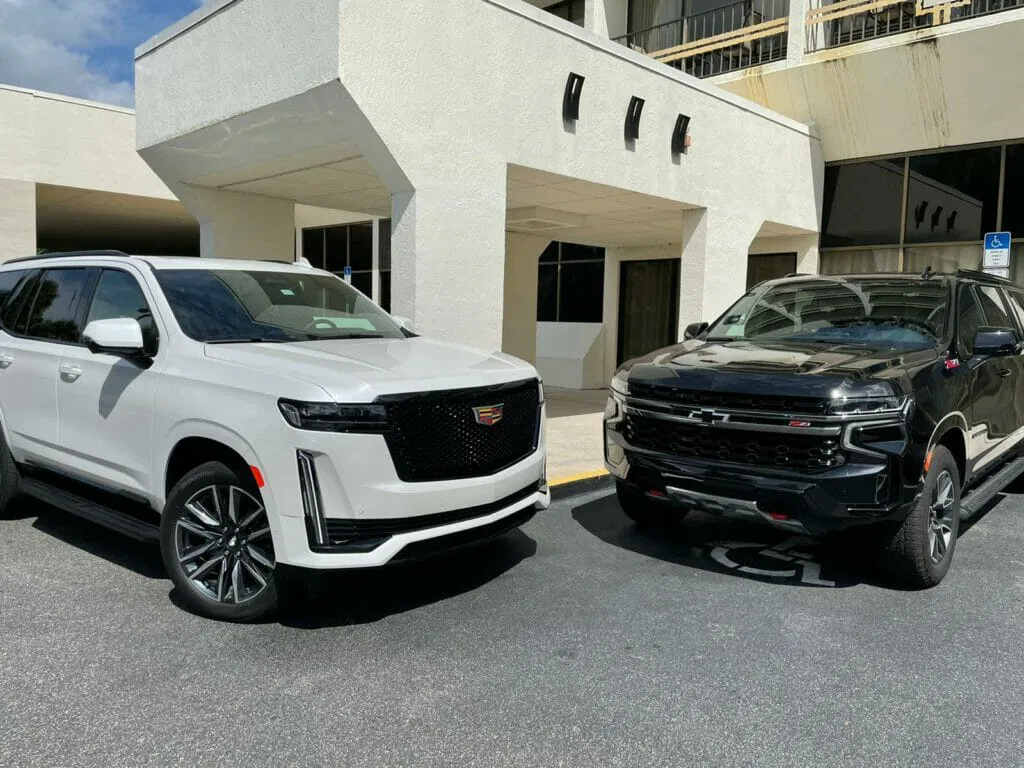 A white Cadillac SUV and a black Chevrolet pickup truck, ideal for group transportation or car service, are parked side by side in front of a modern building.