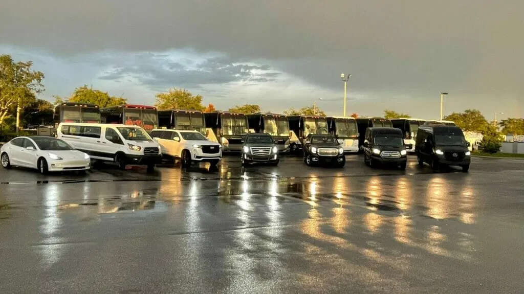 A row of various vehicles—including sedans, vans, SUVs, and buses—ideal for car service or airport transfer, parked on wet pavement under a cloudy sky.