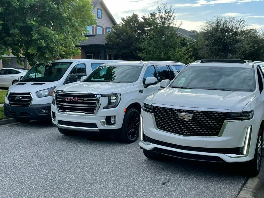 A white Ford van, a white GMC SUV, and a white Cadillac SUV—ideal for group transportation or car service—are parked side by side on a residential street next to trees and houses.