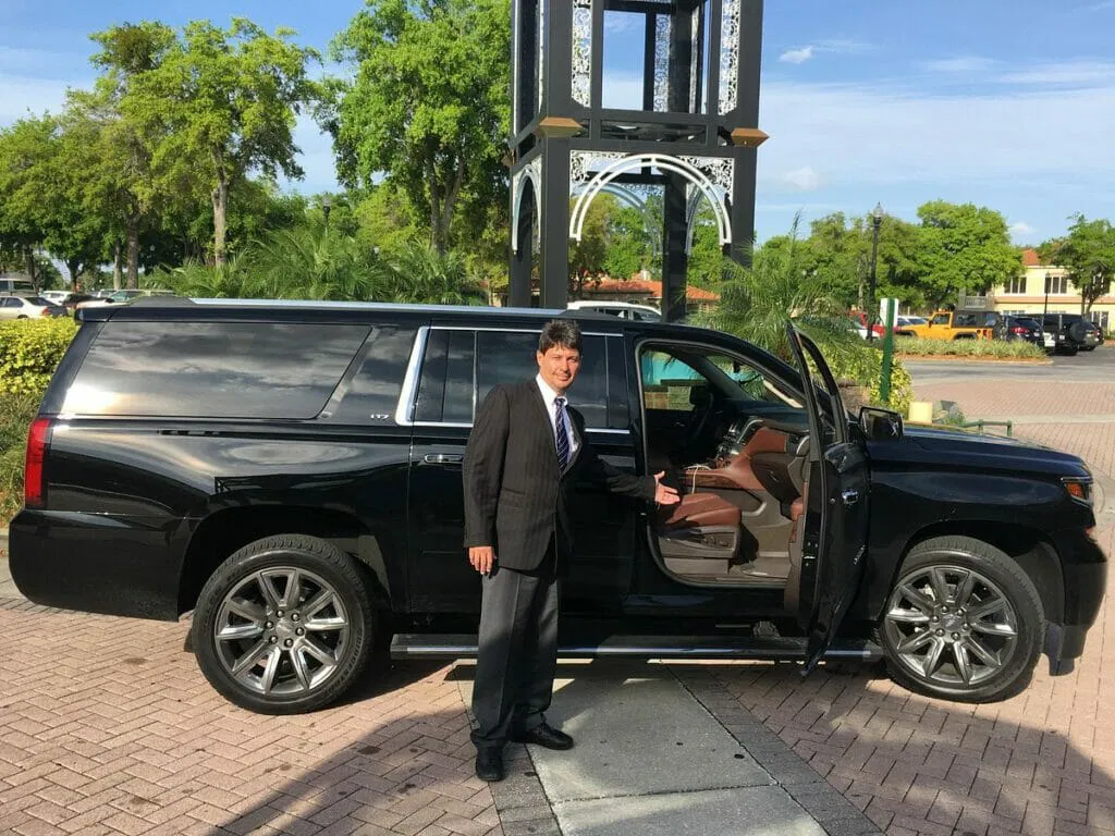 A man in a suit stands next to a black SUV with the driver's door open, gesturing towards the interior—perfect for group transportation or a reliable car service in an outdoor parking area.