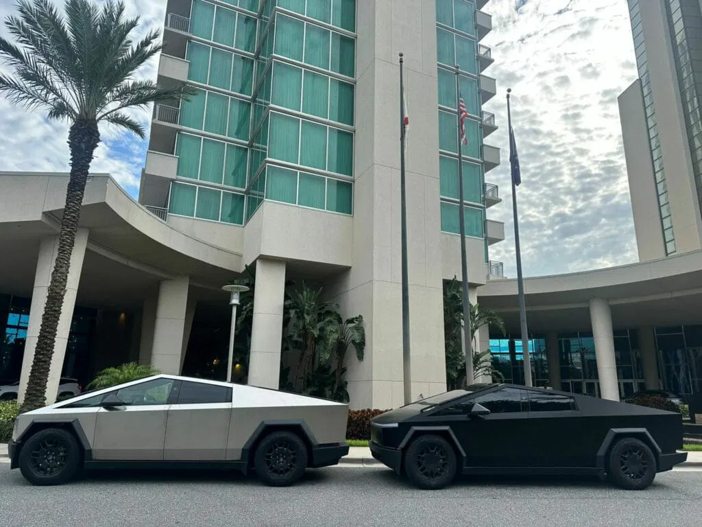 Two Tesla Cybertrucks, one silver and one black, are parked side by side on a street in front of a modern high-rise building with palm trees—perfect for an Orlando airport transfer or luxury car service experience.